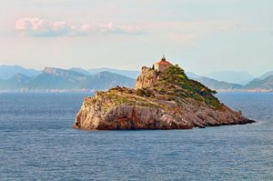 A small island surrounded by a stretch of ocean, hills and mountains in the background. A small house or tower is at the top of the island.
