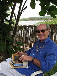 The poet Billy Collins sits in a shady porch overlooking a lake. He’s smiling and holding a stakc of his books. He’s dressed casually, wearing sunglasses and beaded folk-art jewelry.