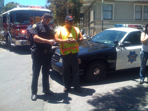 A photograph of two policemen standing on a street corner writing a report. There are a police car and a fire engine in the background.