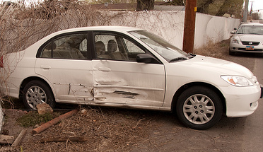 A photograph of a small four door car that has been sideswiped and has crashed into a fence.