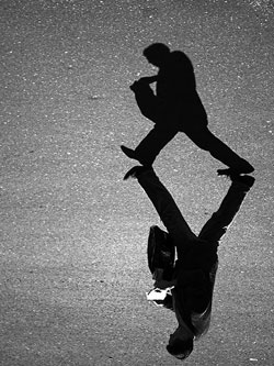 A photograph of a man walking with a notebook in his hand. His shadow appears next to him on the ground.