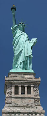 A photograph of the Statue of Liberty. She is a woman wearing robes nad holding a torch aloft. She is holding a tablet and is wearing a crown of spikes.