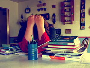 A girl working on English homework holds her head in her arms, obviously very stressed out. Her stack of English textbooks is about eight inches thick.