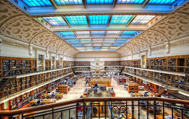 The interior of a vast library. The blue sky above shines down through skylights on a room with thousands of books.