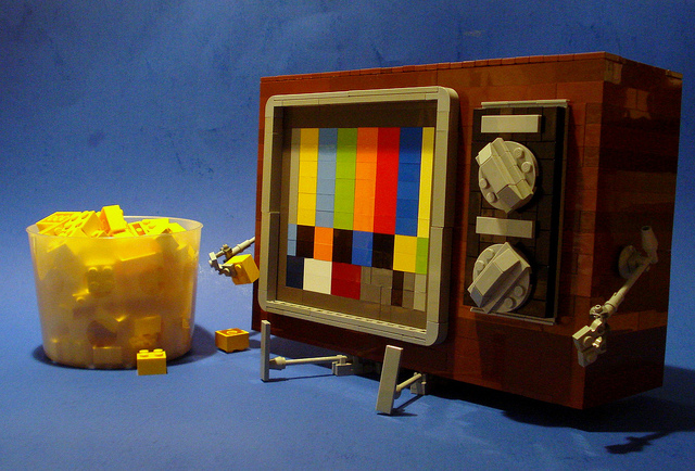 A colorful sculpture made out of legos, showing a television set with arms and legs. The test pattern on its screen resembles a face, and it slowly eats yellow blocks out of a tub, as if it were eating potato chips.