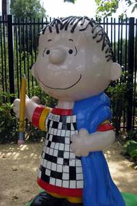 A photograph of a statue of the Peanuts character Linus. He is holding his security blanket and a pencil. On his shirt is a crossword puzzle with his name filled out in one of the sections.