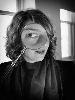 A woman wearing a scarf looks at a camera through an antique magnifying glass as her picture is taken.
