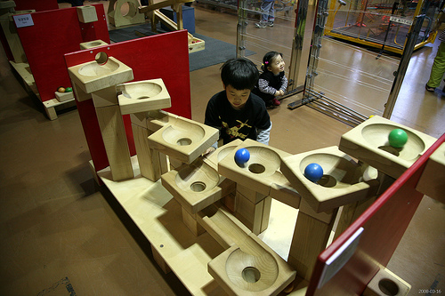 A child in a museum filled with motion sculpture. He watches blue and green balls rolling through a series of funnels.