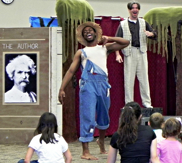 A photograph of a stage production of Huckleberry Finn. There are two actors on the stage in front of an audience of children.