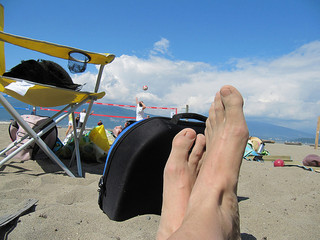A photograph of a beach scene. In the background are people playing beach volleyball and some folding chairs. In the foreground are a person’s crossed feet.