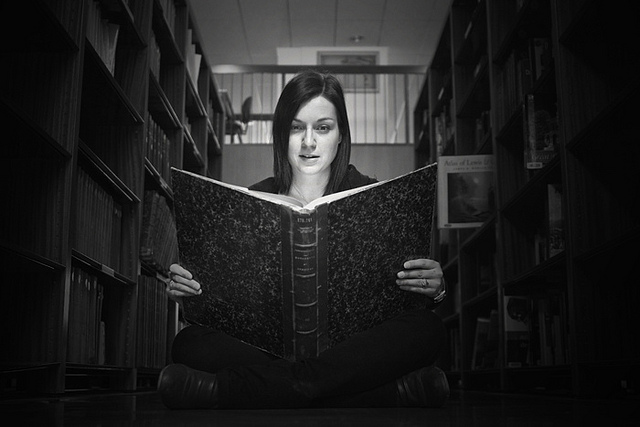 A girl with a huge, old-looking book open on her lap as if reading. She sits between two high shelves staring as if at the viewer of the picture.