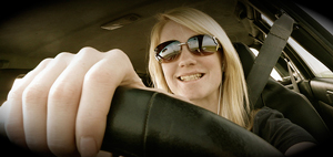 A photograph of a girl driving a car, wearing sunglasses, and grinning.