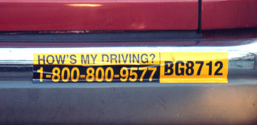 Close-up of a car's bumper sticker; it reads “HOW’S MY DRIVING? 1-800-800-9577 BG8712.”