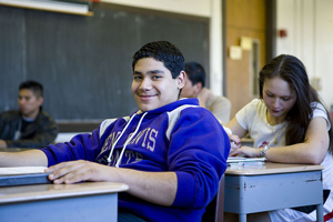 = A photograph of several Latino students seated in a classroom. One, a male student, is looking at the camera and smiling.