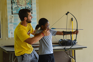 A photograph of a young adult using a bow and arrow while being assisted by an adult male.