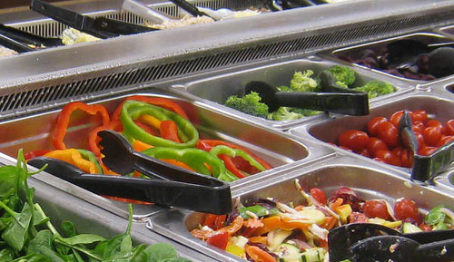 A photograph of a salad bar in a cafeteria. Shown are containers of Bell Peppers, Cherry Tomatoes, Broccoli, and mixed vegetables.