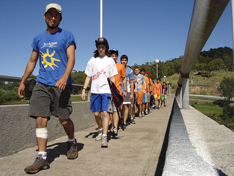 A photograph of a group of teenagers walking across a bridge in a single file line