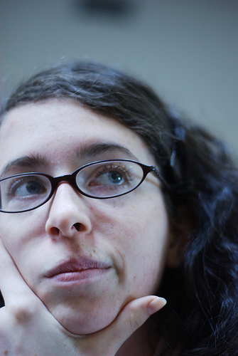 A photograph of a girl wearing glasses holding her chin like she is deep in thought.