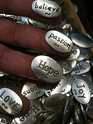 Fingers running through a box of silvery coin-like tokens, each with a word like “hope,” “passion,” or “believe” written on it.