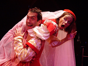 close up of two actors playing Kate and Petruchio from Taming of the Shrew. A jovial Petruchio carries a protesting Kate across one shoulder. Their costumes are red, pink, and gold.