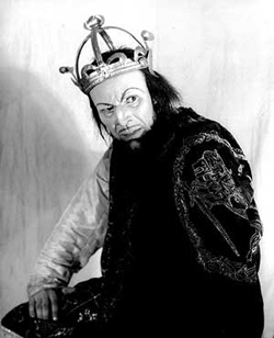 A photograph portrait of an actor dressed as MacBeth. He is wearing a crown and particularly evil looking.