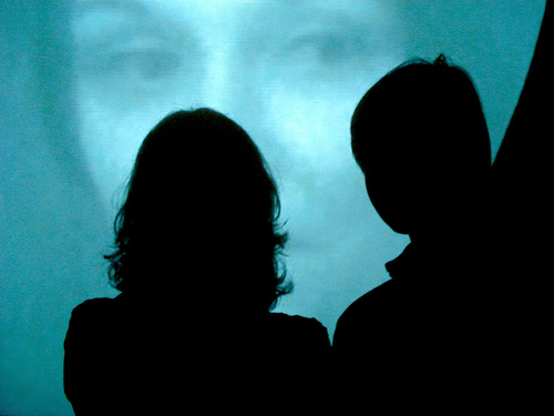 A photograph of two people watching or looking at something; They are silhouetted against the screen.