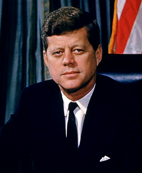 A photograph portrait of President John F. Kennedy. He is wearing a suit and sitting in front of an American flag.