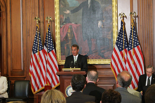 A photograph of Speaker of the House John Boehner addressing an audience in a room. He is flanked by American flags and is speaking from behind a podium.