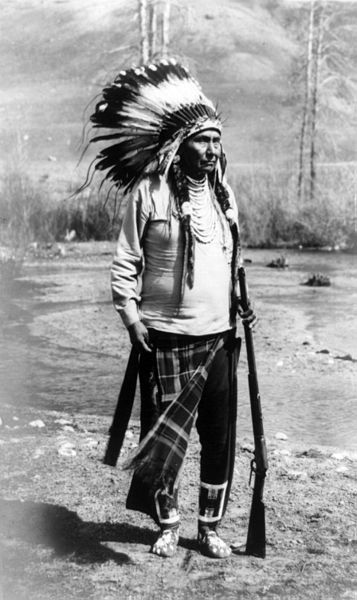 A photograph of Chief Joseph of the Nez Perce tribe taken in 1903. He is an older man wearing a feathered war bonnet and other American Indian garments. He is also holding a rifle.