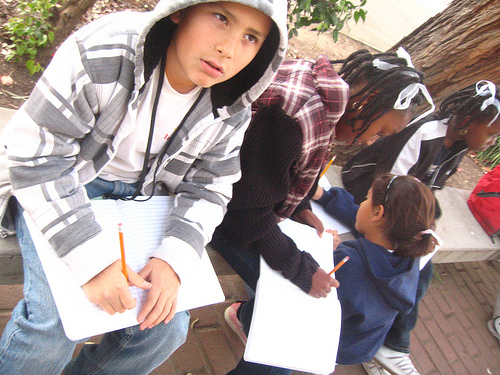 A photograph of a several young students sitting on a prak bench writing in notebooks