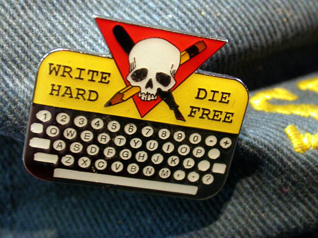 lapel pin containing a keyboard and, above it, a pencil and pen crossing a skull; the words “write hard” and “die free” on either side of the skull.