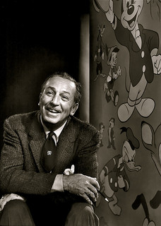 A photograph of animation pioneer Walt Disney. He is seated next to a mural depicting some of his more famous characters: Daffy Duck and Mickey Mouse.