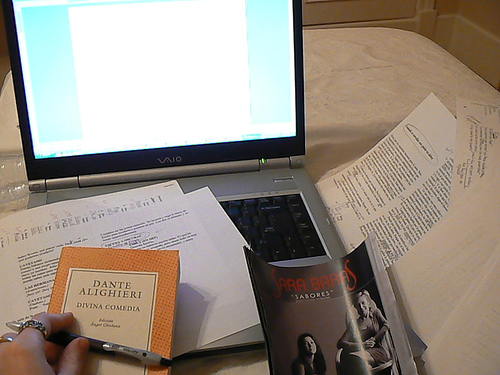 A photograph of a desk with an open laptop computer, written notes and several books on it. There is a female hand holding a pen visible.