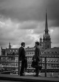 A photograph of two men standing on a bridge having a conversation. There is a cathedral in the background.