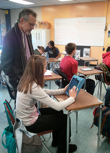 A photograph of a male teacher in s a classroom looking at a student’s laptop or tablet.