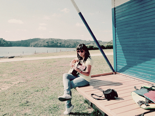 Sitting on the back porch and playing a guitar. A young woman wearing heart-shaped glasses plays, in the background a wide field, forests in the distance. The image is serene and quiet.