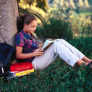 Girl seated outside reading, with school books beside her.
