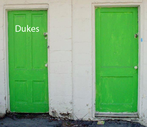 A photograph of two green bathroom doors at an abandoned convenience store.  The doors have padlocks on them.  One door has the word 'Dukes' on it.