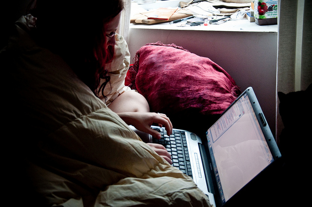 A photograph of a teen aged girl sitting in bed typing on a laptop computer