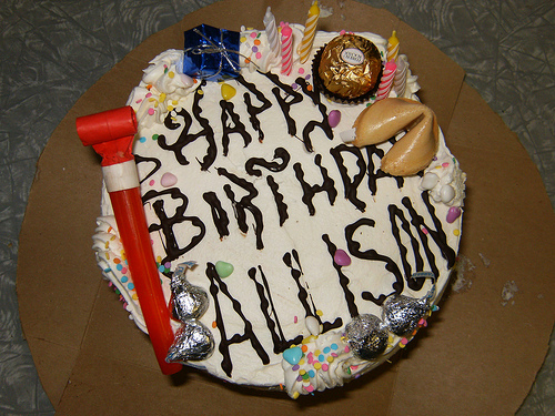 A photograph of a birthday cake that has “Happy Birthday Allison” on it, along with several candies, party items, and a fortune cookie.