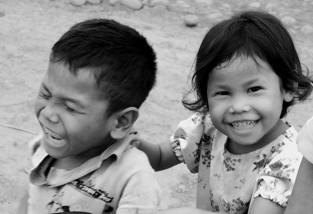 A photograph of a brother and sister. Both are under 4 years old. The sister is smiling and the brother is upset and screaming. He may be laughing.
