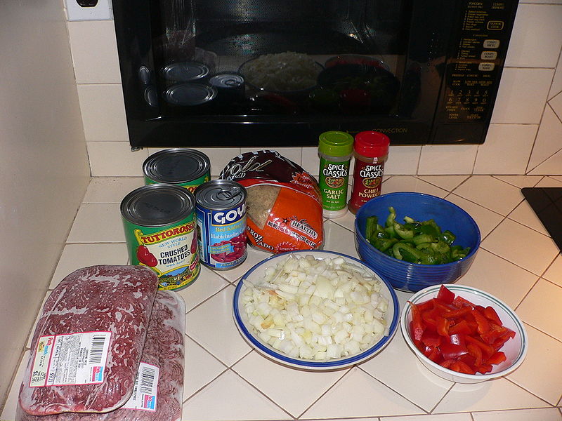 A photograph of chili ingredients on a kitchen counter. Shown are ground beef, cans of various beans, a bag of rice, containers of garlic salt and chili powder, cut & chopped jalapenos, onions, tomatoes.