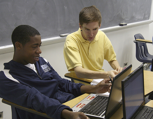 A photograph of a male teacher working with a male student. The teacher is looking at and pointing to something on the student’s laptop computer screen.