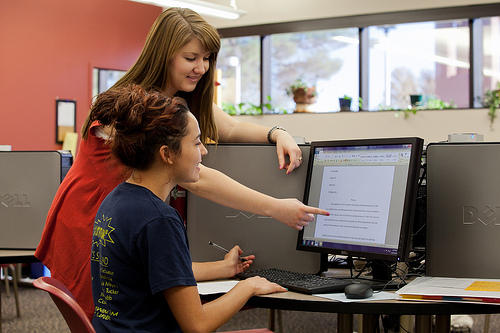 A photograph of a female student and a female teacher working together. The teacher is pointing to something on the computer screen and the student is writing something down in a notebook.
