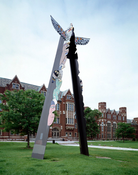 A photo of a modern version of a totem pole that is constructed of metal and stands on a lawn in front of several red brick buildings; animal motifs appear on the totem pole