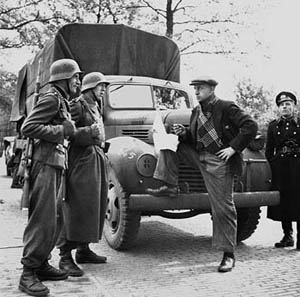 A photograph of several World War II era German soldiers standing next to a large truck. The soldiers are talking to man dressed in civilian clothes.