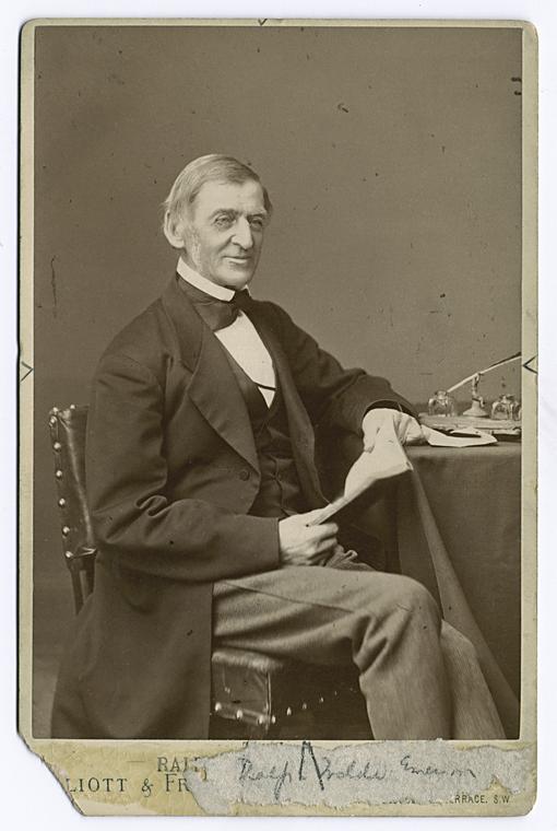 A photograph of Ralph Waldo Emerson sitting at a desk. He is a man in late middle age wearing a suit and tie typical of the 19th century.