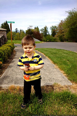 A photograph of a little boy standing on the grass at the edge of where a sidewalk has ended
