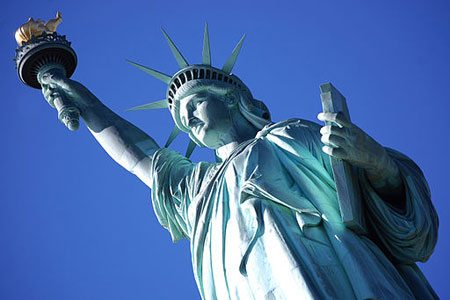 A photograph of the Statue of Liberty. She is dressed in a toga, holding a tablet and hoisting a torch. Her head is crowned with a spiked crown.