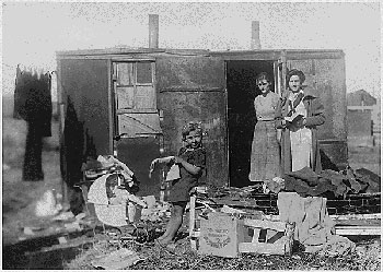 Two women and a girl child stand outside a tatterdemalion, box-shaped tenement; haphazard elements of homemaking fill the bottom of the frame